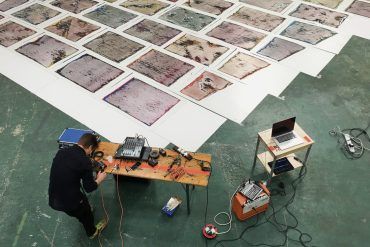 Idle Chatter, overhead view of large room with painted tile artworks on the floor, table with experimental electronic music gear, musician