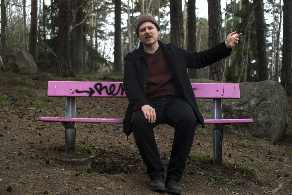 John Chantler sat on a pink park bench surrounded by forest, pointing behind him