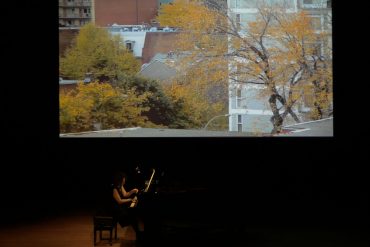 Eve Egoyan playing a grand piano with a projected image of autumn trees and buildings behind her