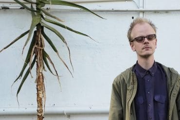 Johan Lindvall - solo/ensemble, composer with his eyes closed stood next to a tall tropical plant