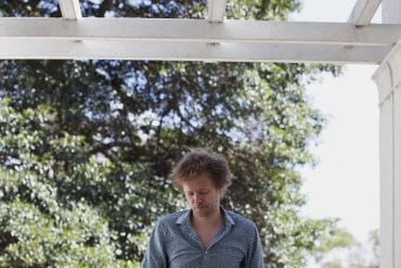 Kim Myhr - Bloom, artist beneath a white wooden frame structure, green trees in the background