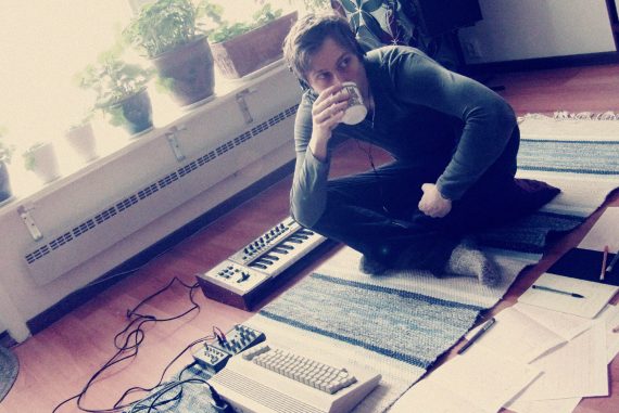 Marcus Fjellström sat on a rug in a living room drinking from a mug, surrounded by audio equipment, notebooks, and indoor plants