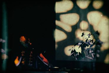 Mytrip performing in a dark room with a small tree and shadows on the wall behind him, photo by Michal Bukolt.