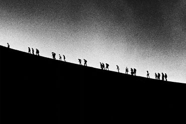 Scott Worthington - Orbit, silhoettes of people climbing a hill against a big grey sky.